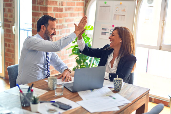 Zwei Personen in Businesskleidung geben sich in einem Büro mit Laptop und Unterlagen am Tisch einen High-Five.