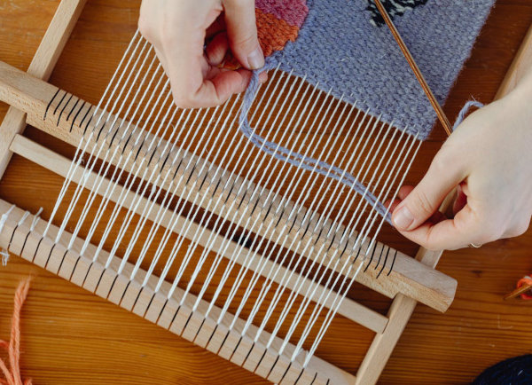 Hände weben mit grauer Wolle auf einem Holzwebrahmen, bunte Wollstücke liegen daneben.