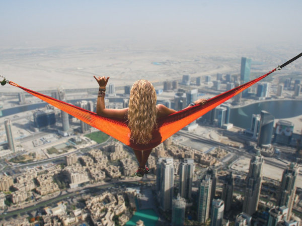 Frau mit langen blonden Haaren sitzt in einer roten Hängematte hoch über einer Stadt mit Wolkenkratzern und Wüstenlandschaft im Hintergrund.