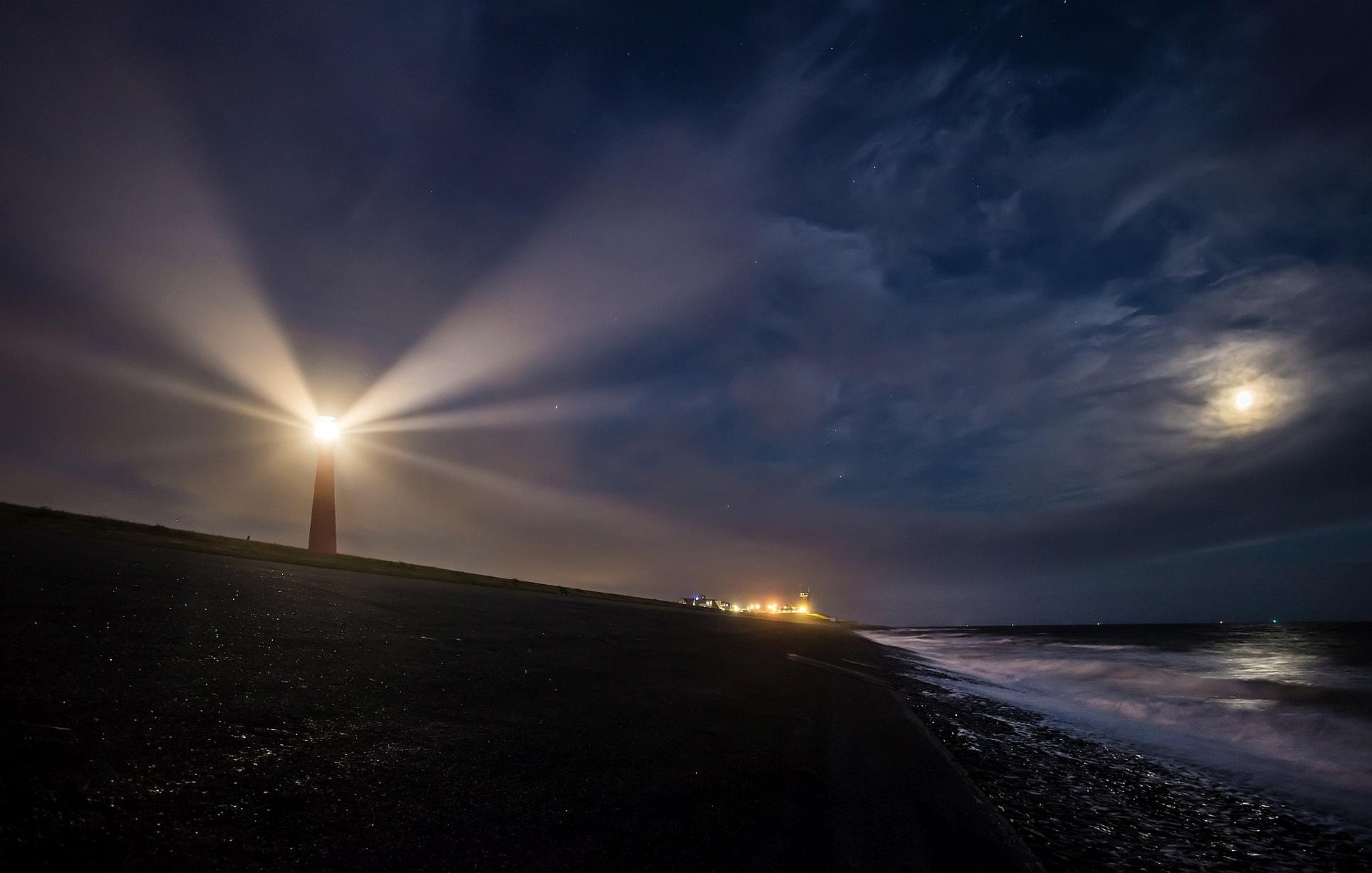Leuchtturm mit hellem Lichtstrahl bei Nacht an dunklem Strand, rechts Meer mit Wellen und Mond am bewölkten Himmel.