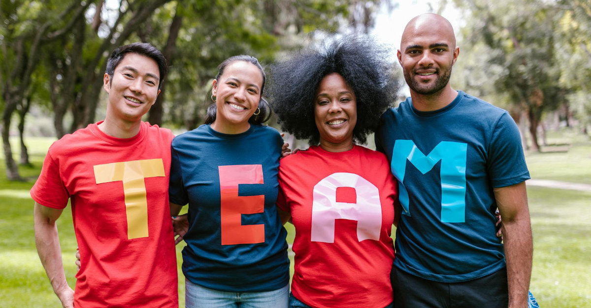 Vier Personen stehen nebeneinander im Freien, tragen bunte T-Shirts mit den Buchstaben 'T', 'E', 'A' und 'M' in großen, kontrastierenden Farben.