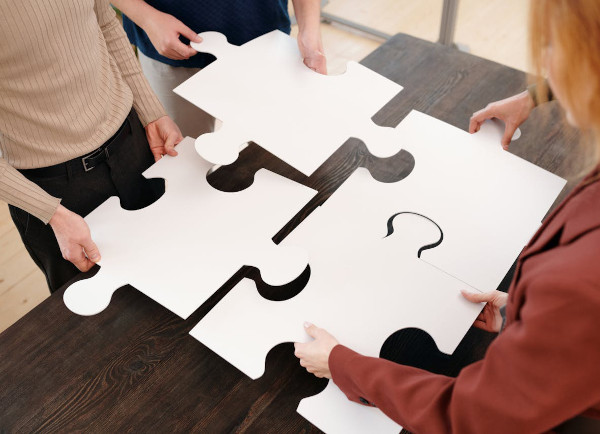 Vier Personen setzen vier große weiße Puzzleteile mit Fragezeichen zusammen auf einem Holztisch zusammen.