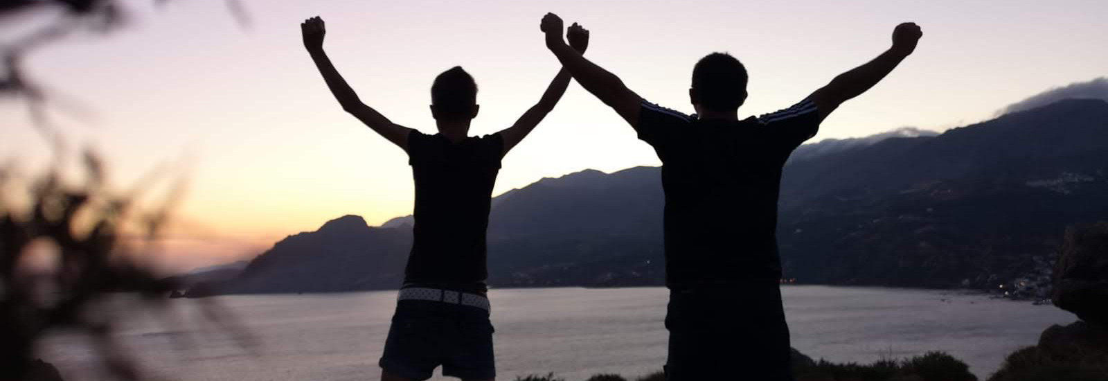 Zwei Personen stehen mit erhobenen Armen vor dem Meer und Bergen im Sonnenuntergang.