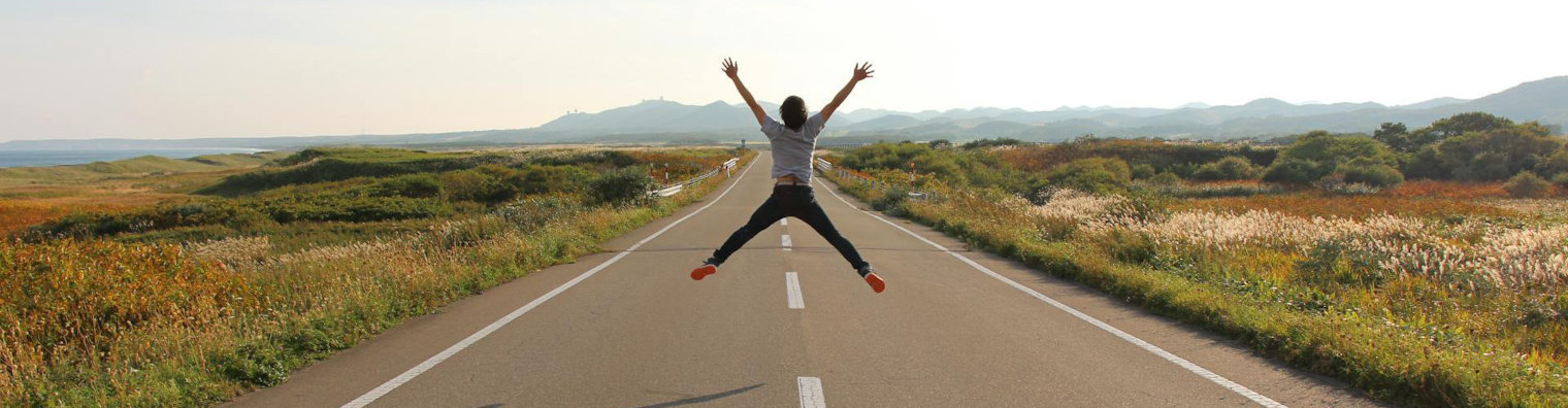 Person springt mit ausgestreckten Armen und Beinen auf einer leeren Landstraße, umgeben von Feldern und Bergen im Hintergrund.