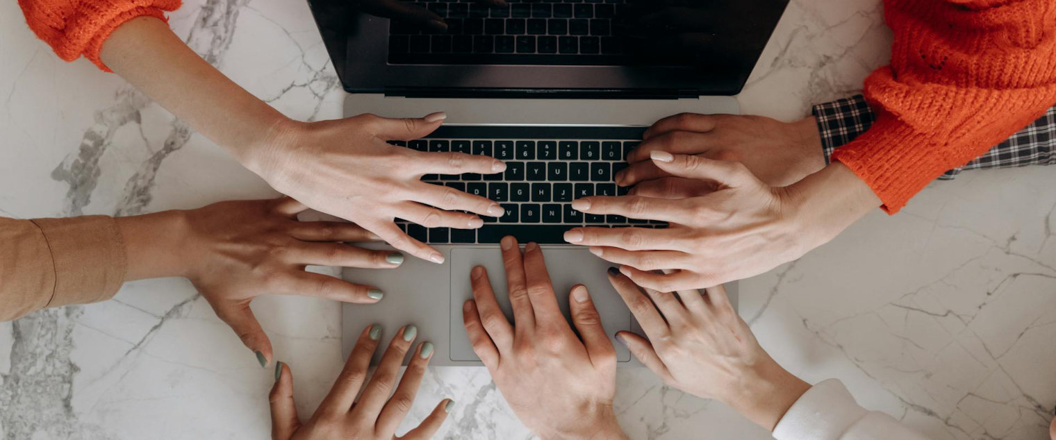 Mehrere Hände unterschiedlicher Personen tippen gemeinsam auf der Tastatur eines Laptops auf einer Marmoroberfläche.
